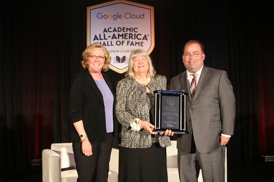 Julie Soriero, Louise Jandura, and Ken Johnson stand on a stage with a black backdrop. Jandura holds a plaque, and a sign behind them reads “Google Cloud Academic All-America Hall of Fame"