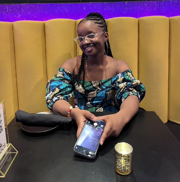 A young Black woman wearing a brightly patterned top and braids smiles over a set table at a restaurant. 