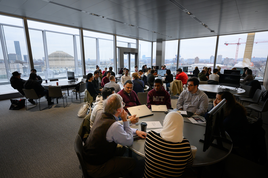 Groups of people sit around around tables in a room surrounded by glass walls with a view of Boston, Cambridge, and of MIT's Great Dome in the background.