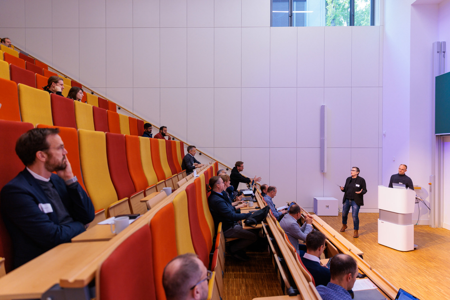 A woman is presenting in front of a small audience in a lecture hall with bright orange and yellow chairs.