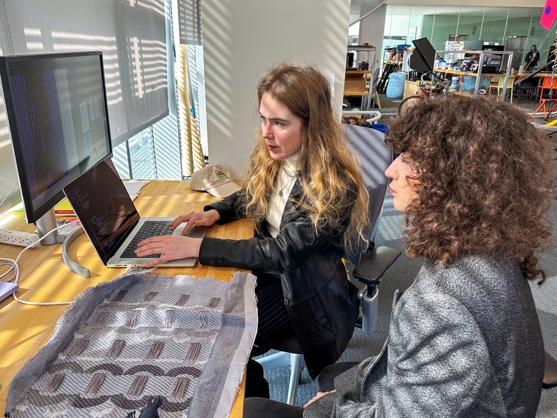 Two women sit at a desk with an open laptop, a large monitor, and a piece of textile. One is typing and talking while the other looks on.
