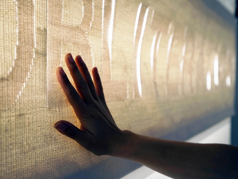 A silhouetted hand touches a long white, backlit, woven textile.