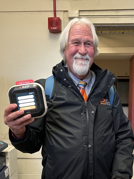 David Barber stands in an MIT office and holds an AED device up to the camera. The device’s touchscreen display is illuminated.