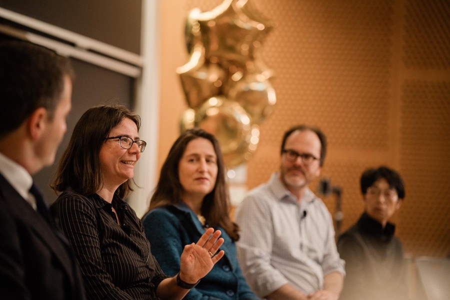 Ana Bell, Melissa Webster, Jesse Thaler, and Takako Aikawa sit in a line in front of a lecture hall. Bell is gesturing and speaking. An unidentified man looking away from the camera sits with them.