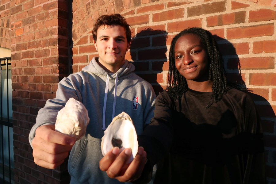 Santiago Borrego and Unyime Usua stand outdoors in front of a brick wall, each holding out an oyster shell.
