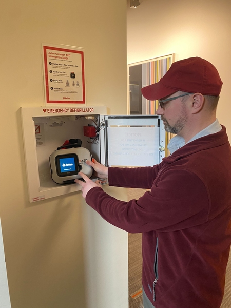 Steve Taddonio operates an AED device that is placed in a wall cabinet in NW23. A sign with operational instructions is displayed on the wall above the cabinet.