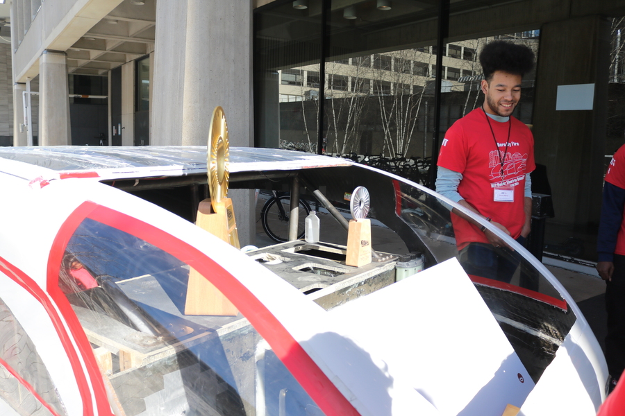 A student wearing a red SEVT shirt smiles behind the solar car, holding two trophies
