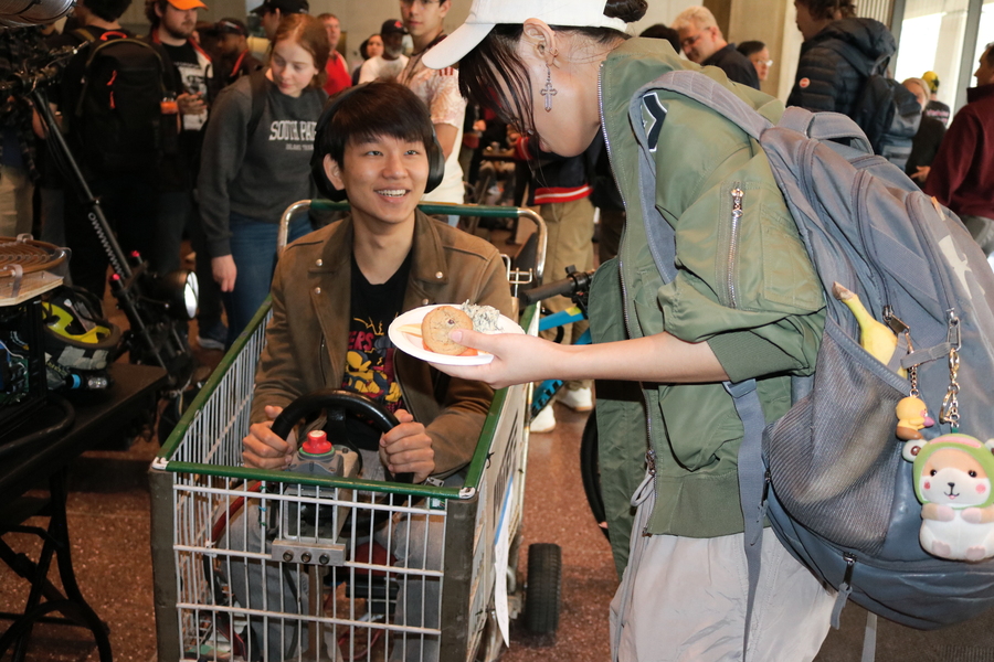 A student holding a plate of food talks to another student sitting in motorized shopping cart