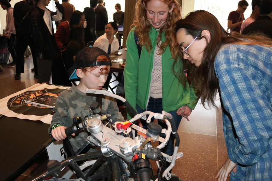 A young child looking at the Electric Vehicle Team's motorcycle and talking to a student