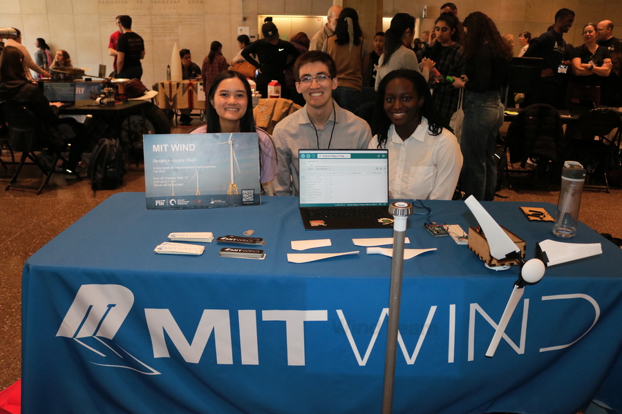 3 MIT students sitting at the MIT Wind Team table, smiling, with their wind turbine prototype 