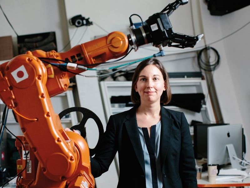 Julie Shah stands next to a large orange robotic arm