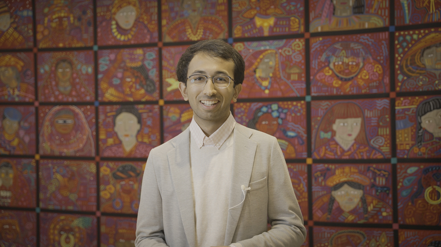 Photo of Arjav Shah standing in from of a colorful mural depicting people in various styles of dress