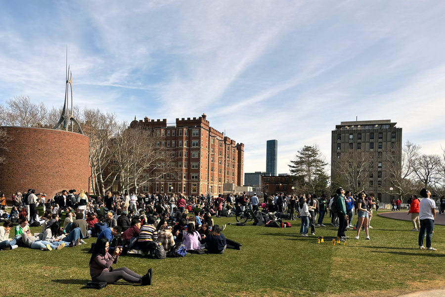 Crowds sit on the grass and hang out, with MIT Chapel and other buildings in background.