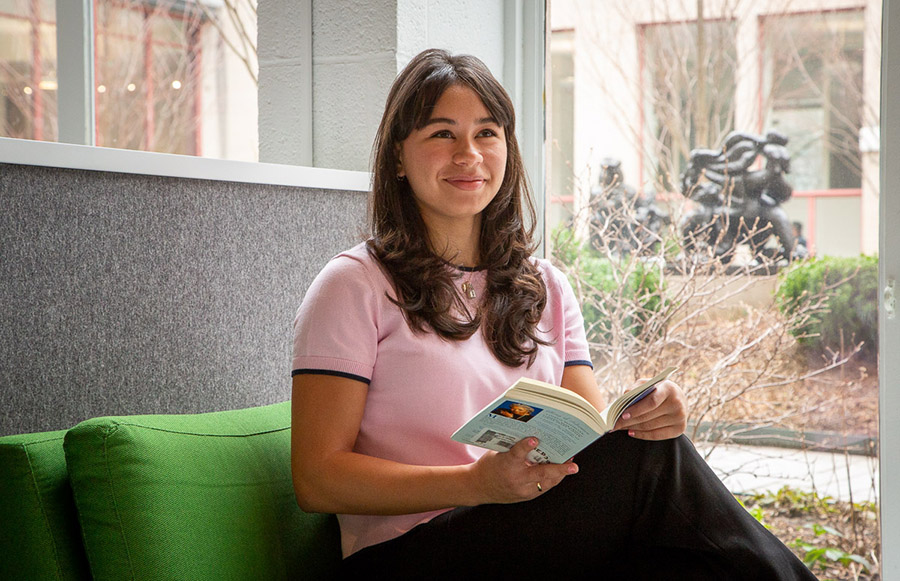 Grace McMillan, holding a book, sits on a low-backed sofa with green cushions. A courtyard is visible through a window behind her.