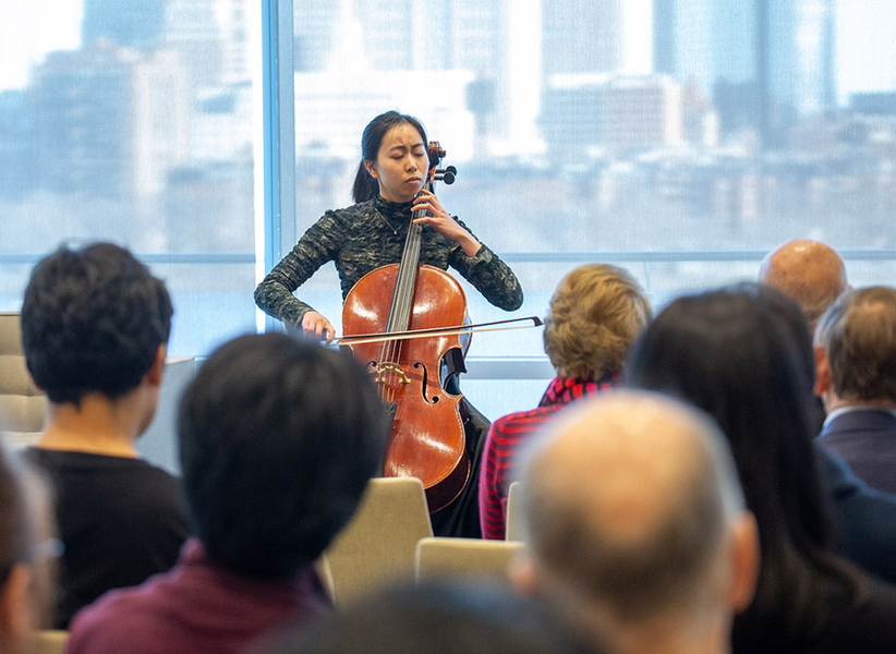 Valerie Chen plays a cello in front of an audience. Behind her are glass windows that look out onto the Charles River and the skyline of Boston.