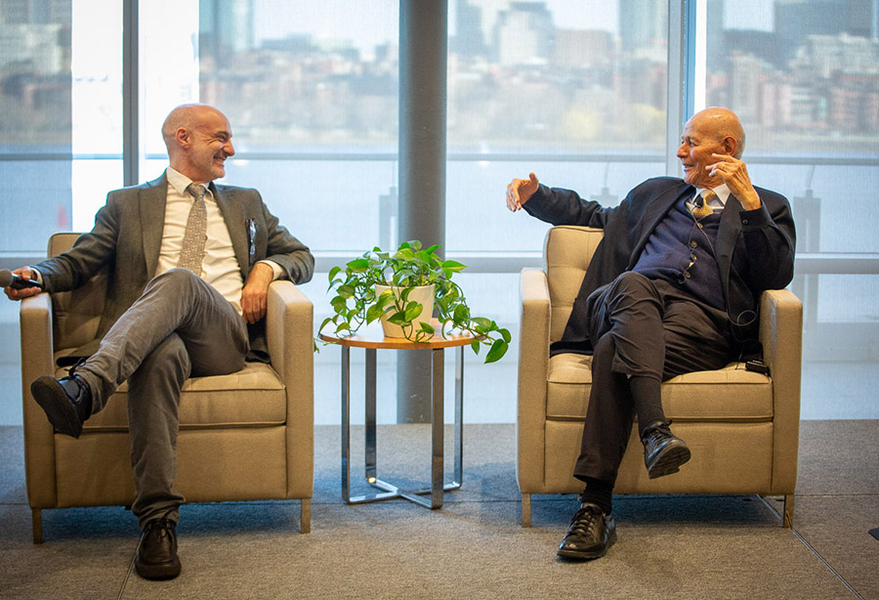 Agustin Rayo and Carlos Priety sit in chairs speaking to each other in front of a set of glass windows that looks out onto the Charles River and the skyline of Boston.