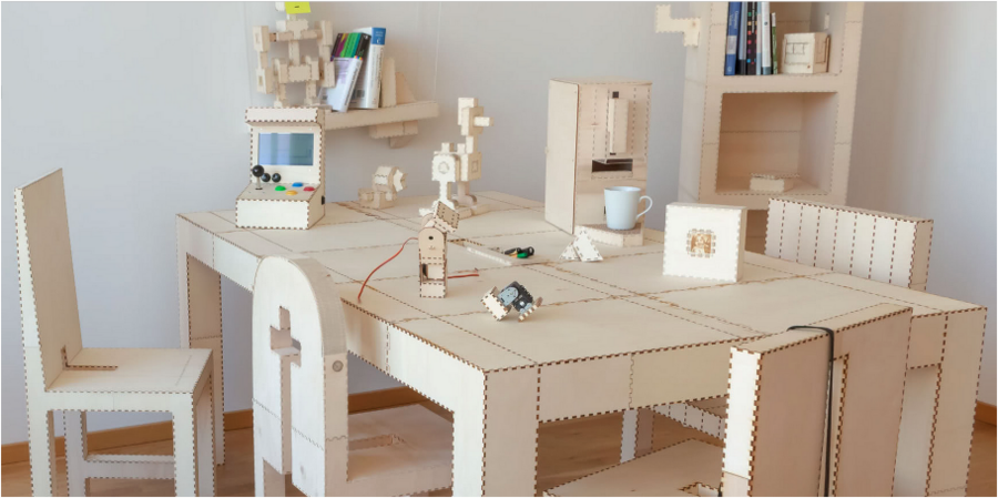 In a small room, life-sized furniture and accessories — table, chairs, shelves, and objects such as a coffee machine, robot, and computer game — are made from thin unfinished wood
