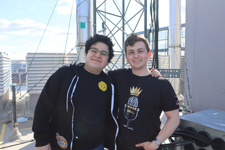 James Rock and Diego Temkin pose on a rooftop with their arms around one another's shoulders. The base of a radio transmitter antenna is behind them.