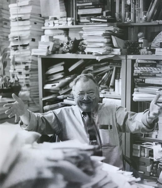 A grayscale photograph of Professor Bernie Wuensch in his office, surrounded by books and heaps of papers, welcoming the camera with open arms and a warm smile
