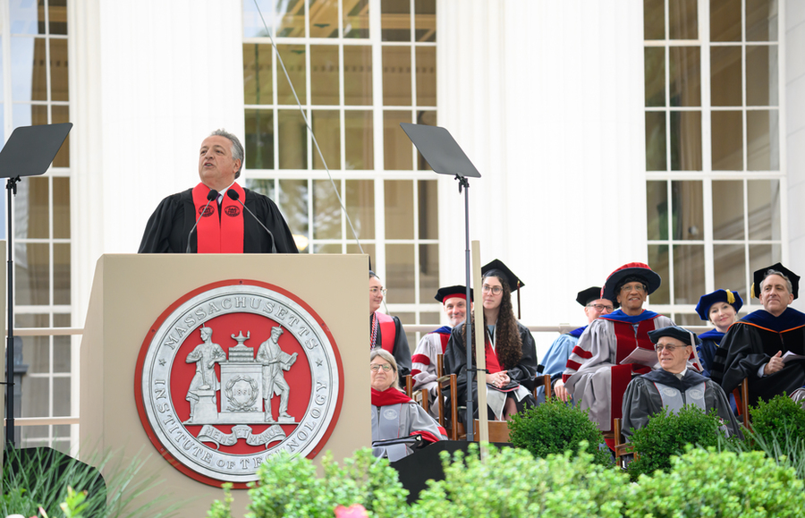 Noubar Afeyan speaks at a podium with the MIT seal on the front. Faculty and administrators in academic regalia are seated next to him.