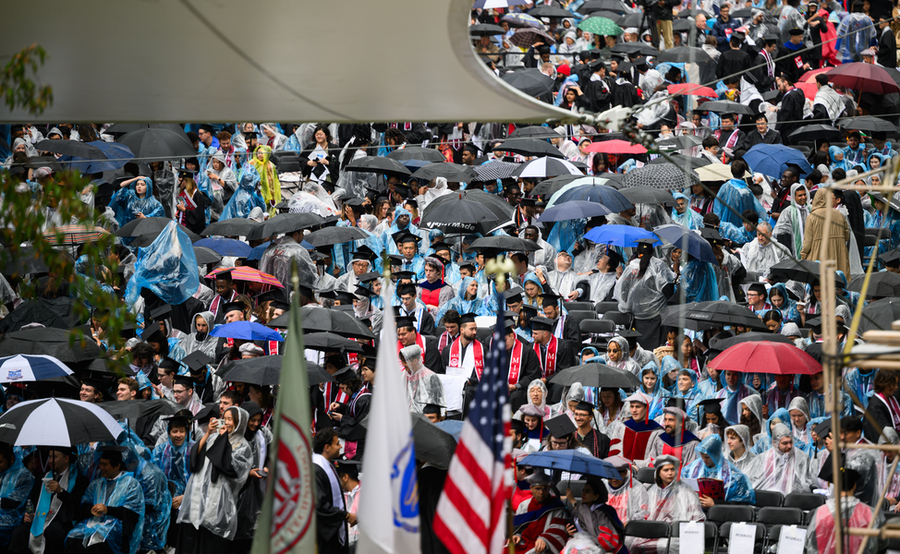 Hundreds of people wearing raincoats and holding umbrella sit in rows on Killian Court.
