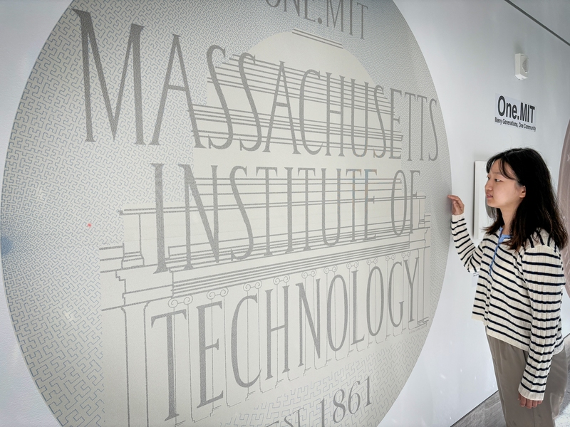 Seungyeon Woo looks at a large circular image on a white wall that includes the words "Massachusetts Institute of Technology," "OneMIT," and a depiction of the MIT Dome.