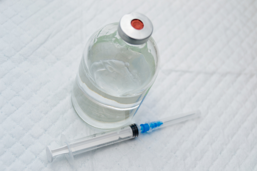 A clear drug vial with a syringe on a white background, seen from above