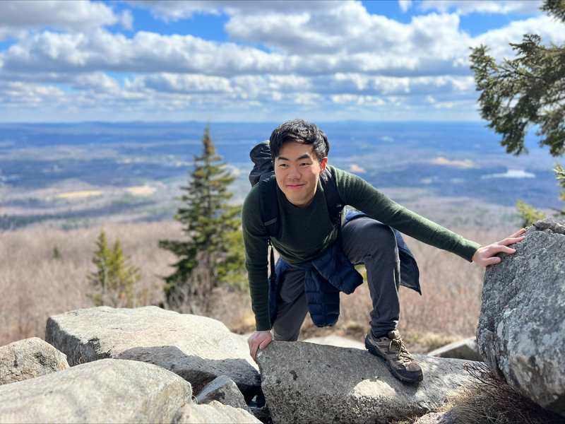 A young man wearing a long-sleeve T-shirt, jeans, and sneakers scrambles over a rocky ledge atop a high mountain. Clouds, a broad sky, and forested hilltops are visible in the background.