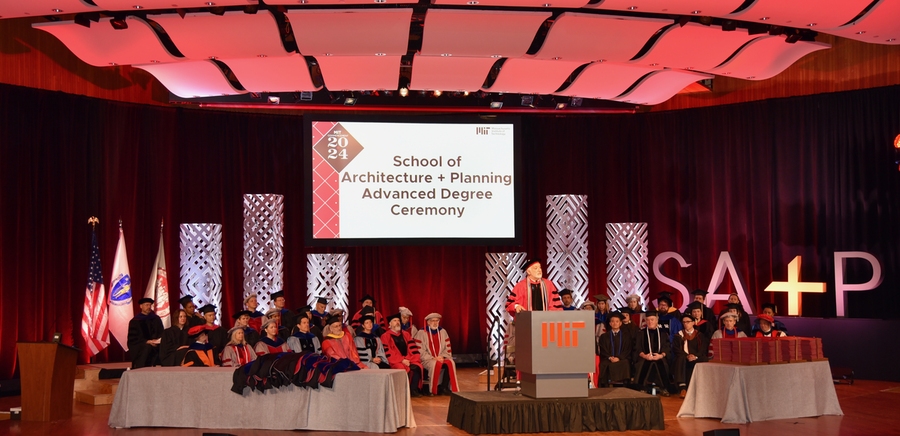 man in an orange graduation gown speaks at a lectern on a stage, with a couple dozen people in ceremonial garb seated behind him.