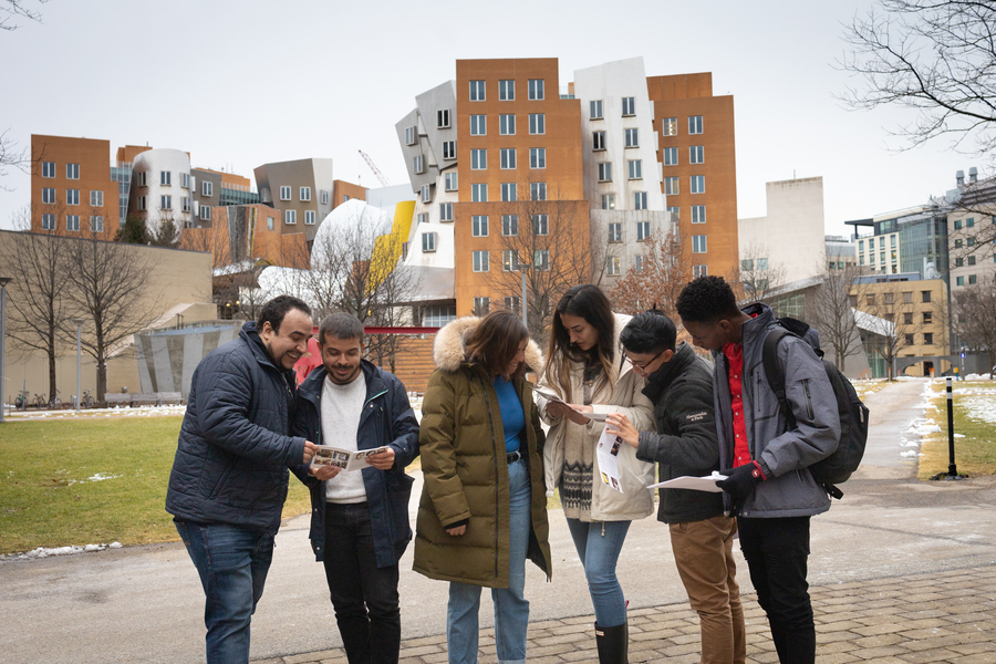 Six students wearing winter jackets consult printed materials and talk with each other in front of the Stata Center, a building of brick and steel with unusual angles