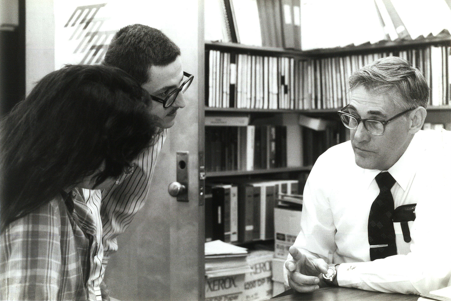 Black and white photo of David Lanning seated and leaning on a table in a room lined with bookshelves filled with books; speaking with one make and one female student standing in a doorway to the room on the left.