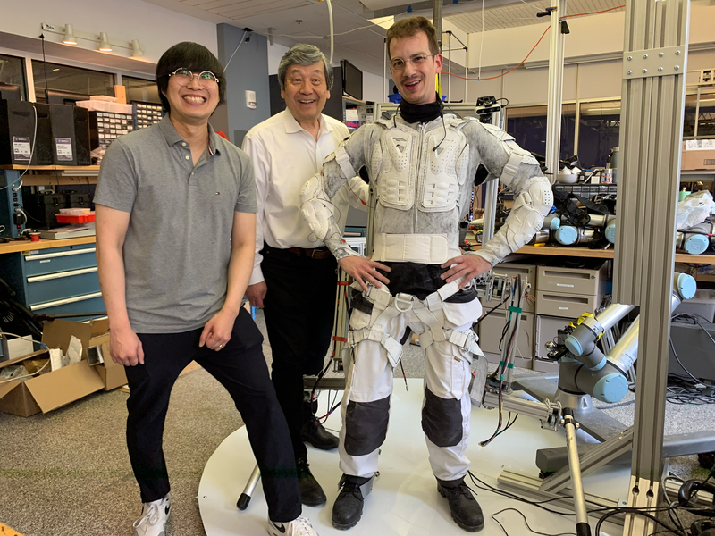 Sang-Yoep Lee, Harry Asada, and Erik Ballesteros stand in a lab. Erik is wearing the inside part of the new robotic suit, which resembles sports padding.