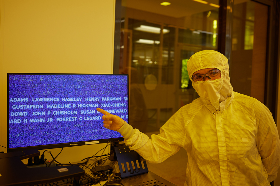 A person in an all-white protective suit stands in yellowish light inside a laboratory pointing to a computer monitor showing a blow-up of names from the One.MIT wafer.