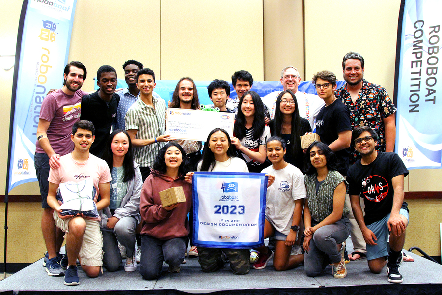 19 people in three rows pose behind a woman holding a sign that reads "First Place Design Documentation."