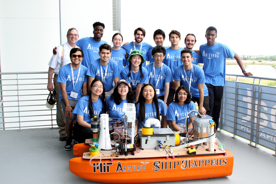 17 people in three rows smile in their team T-shirts, posing behind their orange boat that reads "Ship Happens"
