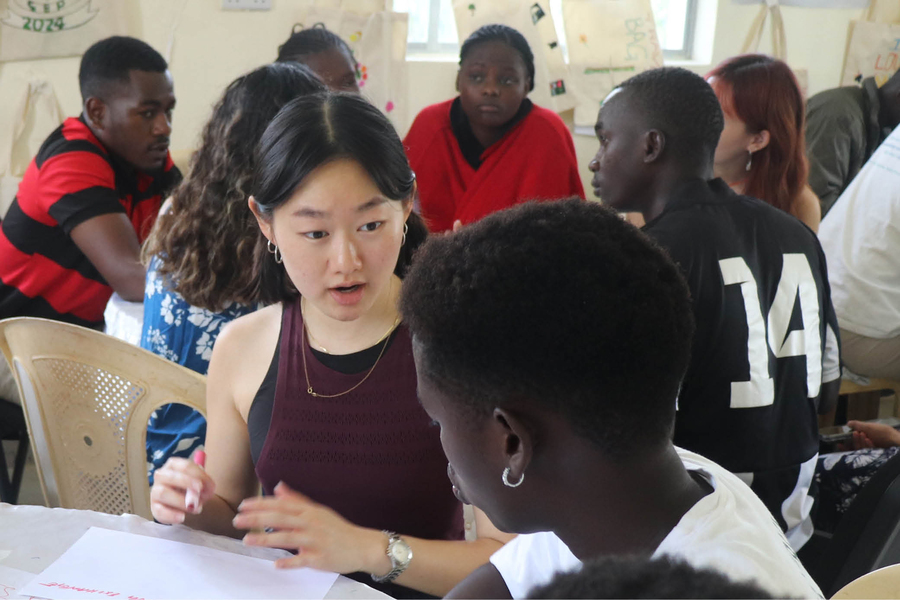 Sophia Chen at a table talking to another woman to her left. In the background is a round table at which are seated four young men and two women.