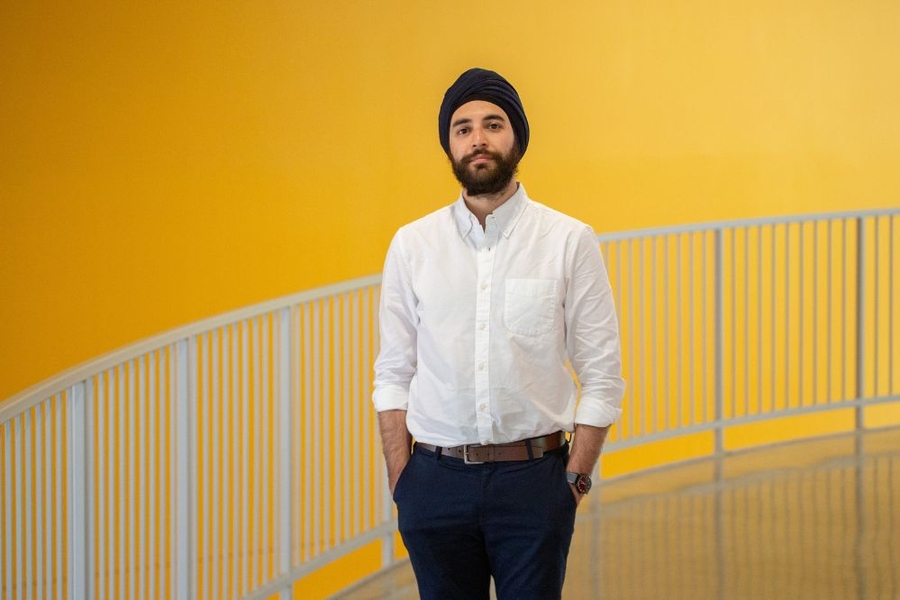 Sukrit Puri, wearing a turban and a white button-down shirt, stands in front of a curved railing against a yellow wall.