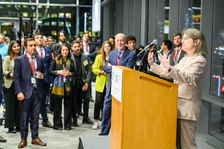 Sally Kornbluth, standing at a lectern, speaks to a standing audience outdoors