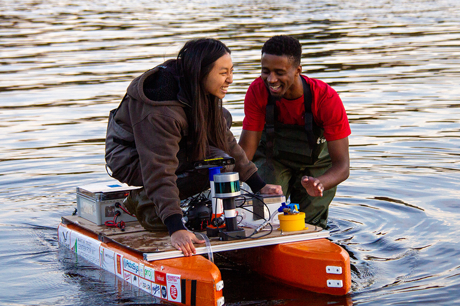 Audrey Chen crouches on a small orange boat that floats on a river while Herbie Turner stands in the water, steadying the boat. Both are laughing.