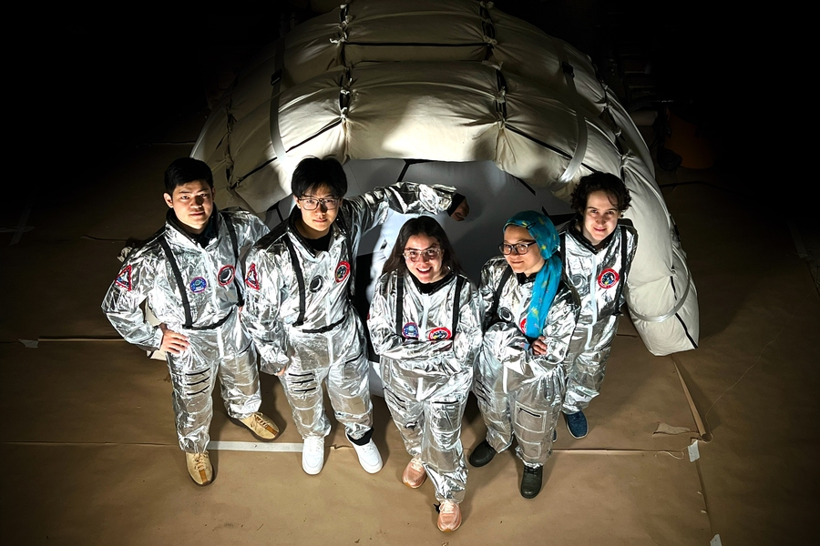 Five people in reflective space gear pose in front of a tent-like structure.