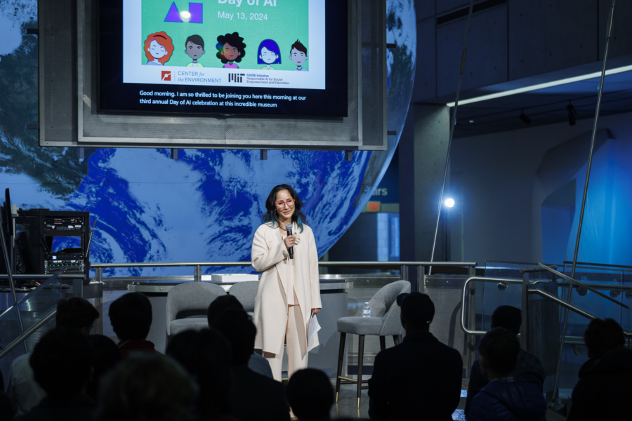Cynthia Breazeal speaks on a stage with a large screen behind her displaying the logos of MIT RAISE and the Center for the Environment. 