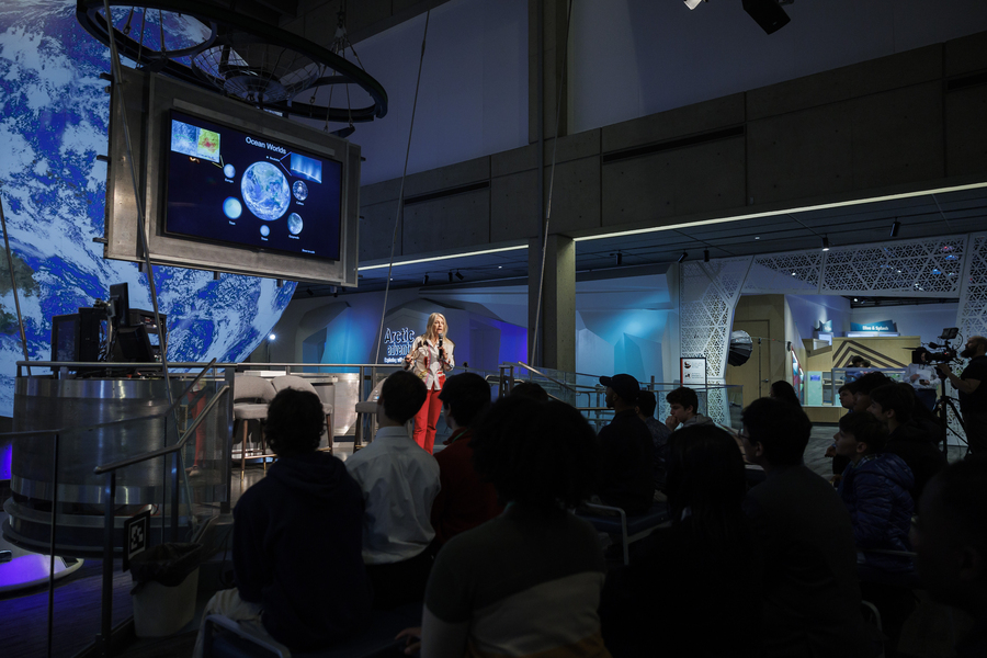 Dava Newman speaks on stage with a large screen displaying images of planets.
