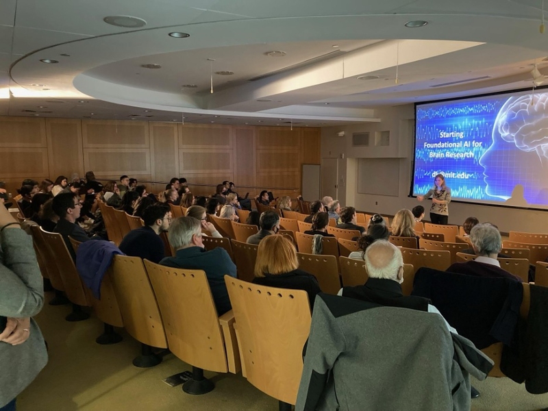 A woman speaks to a packed room of people. A large screen behind her displays a blue slide with the words, "Starting foundational AI for brain research."