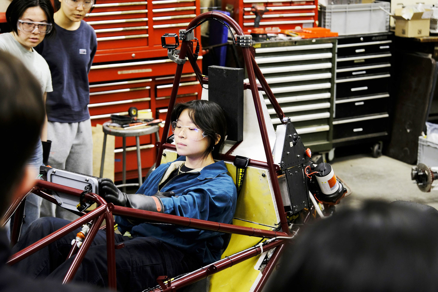 Monica Chan sits in a car frame and reaches up to the dasbboard.