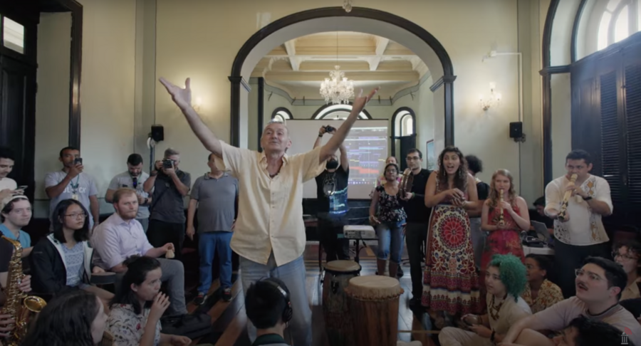 About two dozen MIT students and others gather for a musical experience. A man stands with arms raised in the center of the room.