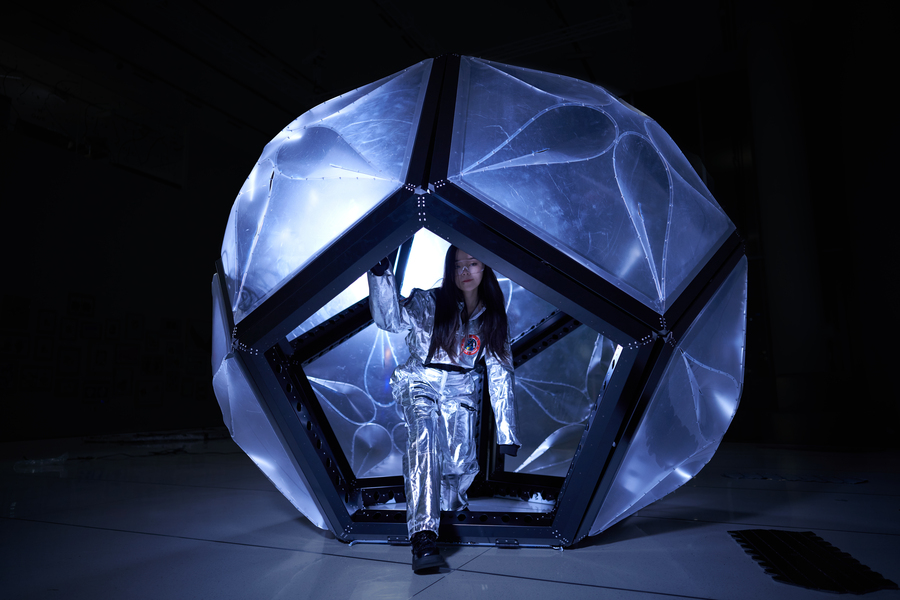 A student in a silver spacesuit steps out of the Momo habitat, a geodesic dome-like structure. 