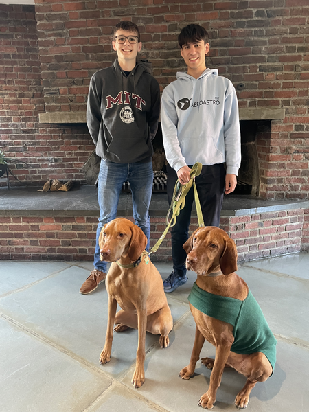 Two students pose in front of a brick fireplace. One has two dogs on leashes.