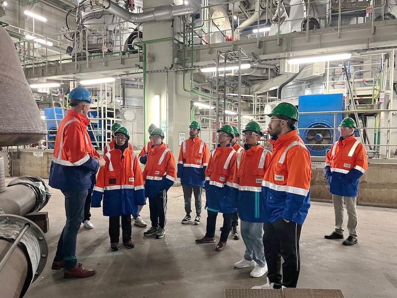 About nine students listen to a man speak inside of power plant. Everyone wears hardhats and safety gear.