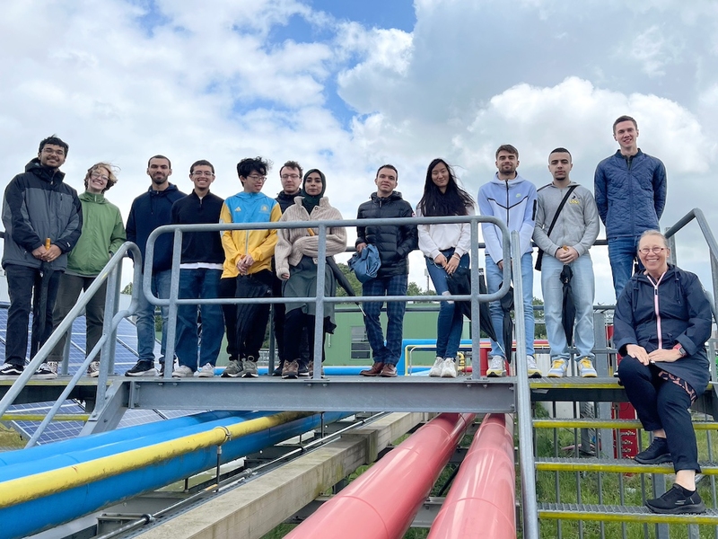 13 people pose together on a catwalk over hydrogen pipelines
