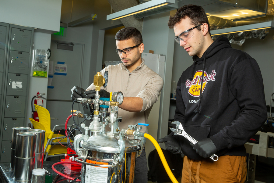 Aly Kombargi and Niko Tsakiris are in the lab wearing safety googles and working with tabletop device with many tubes and valves.